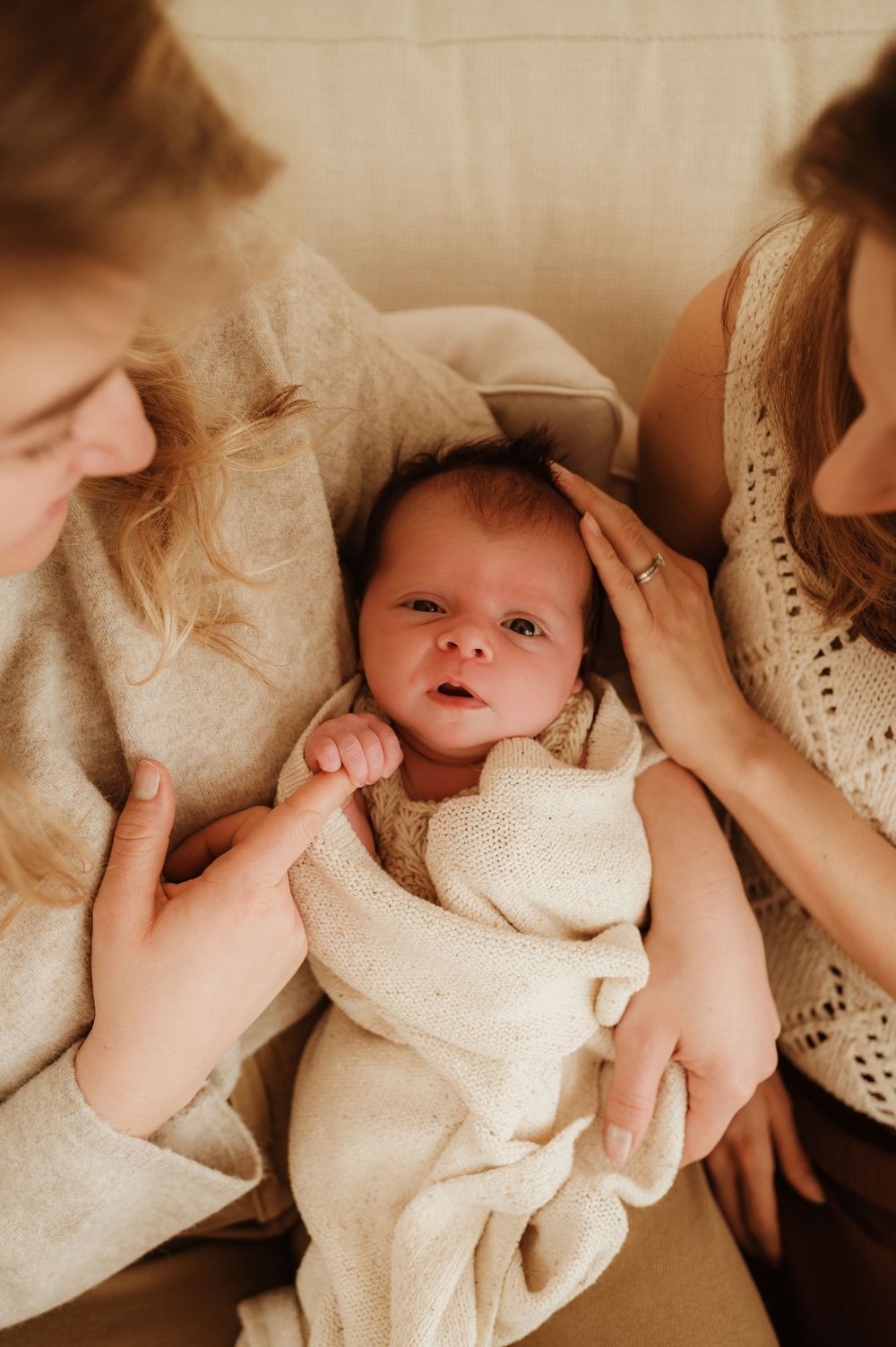 A baby wrapped in cream-colored blankets lies between two people wearing neutral tones in an intimate indoor setting.