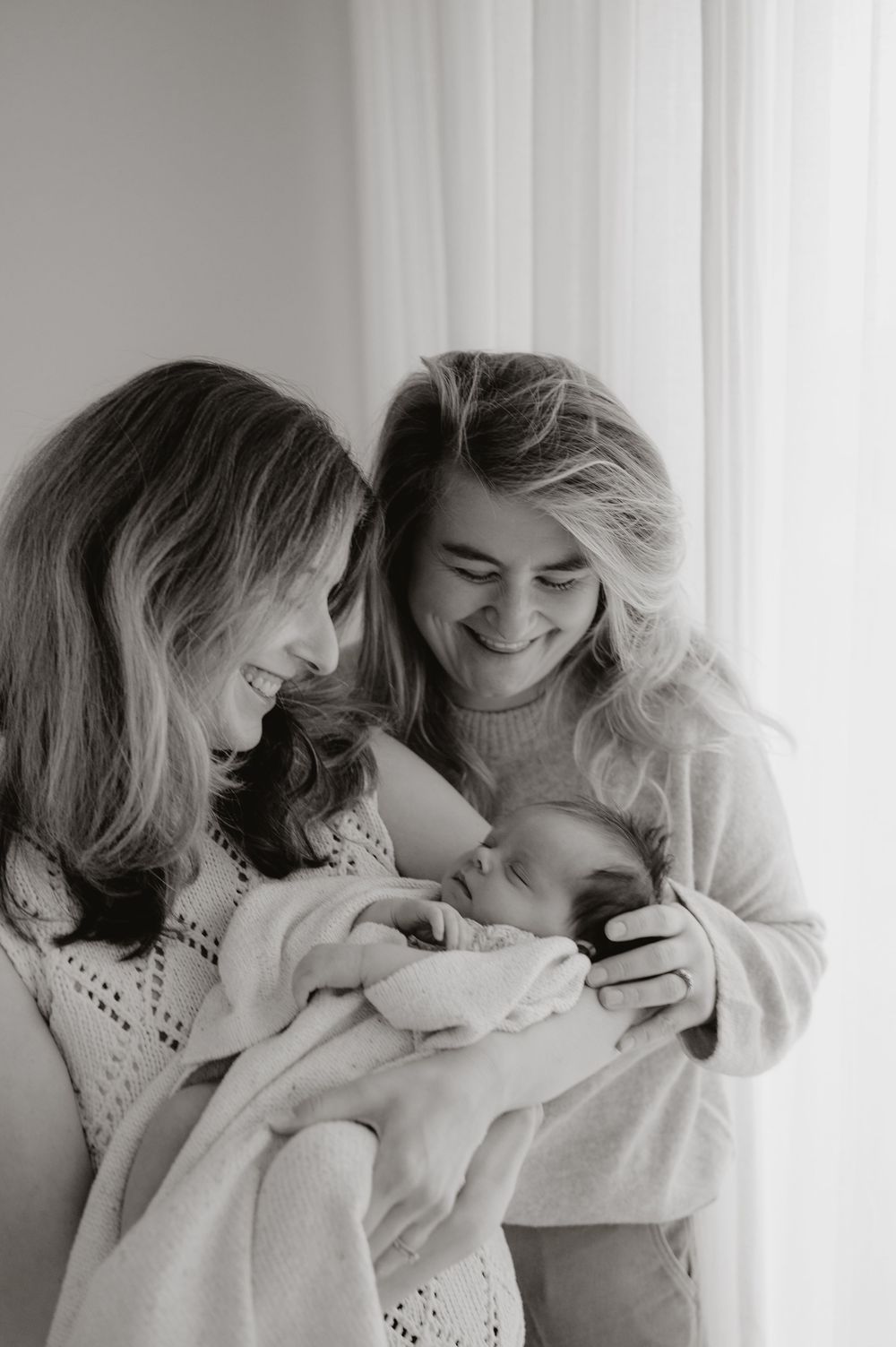 Black and white photo showing two people joyfully holding and gazing at a sleeping baby wrapped in blankets.