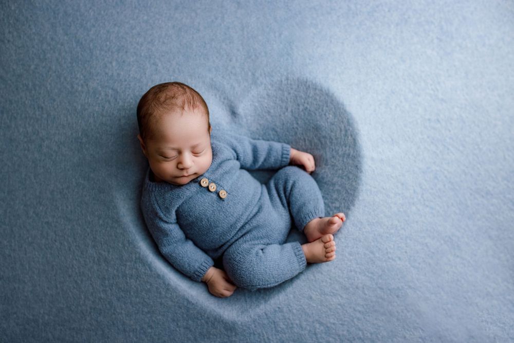 Baby wearing blue sleeper outfit rests on soft blue blanket during newborn photo session.
