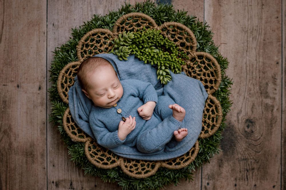 Sleeping baby in blue outfit nestled in decorative green wreath with braided trim on wooden backdrop.