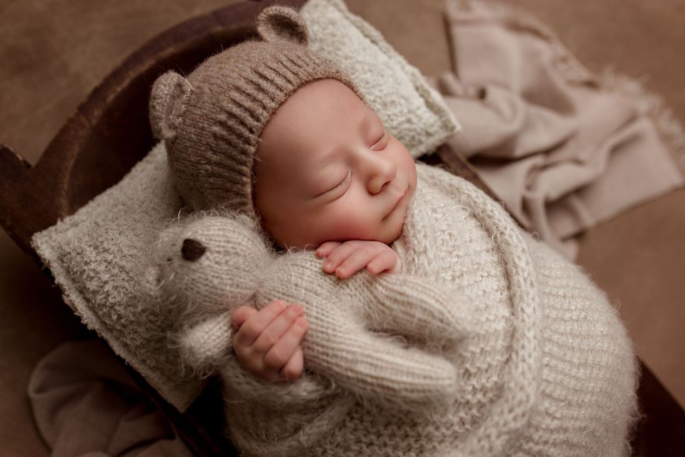 A sleeping newborn wrapped in cozy knit blankets and hat cuddling a small stuffed toy on neutral fabric.