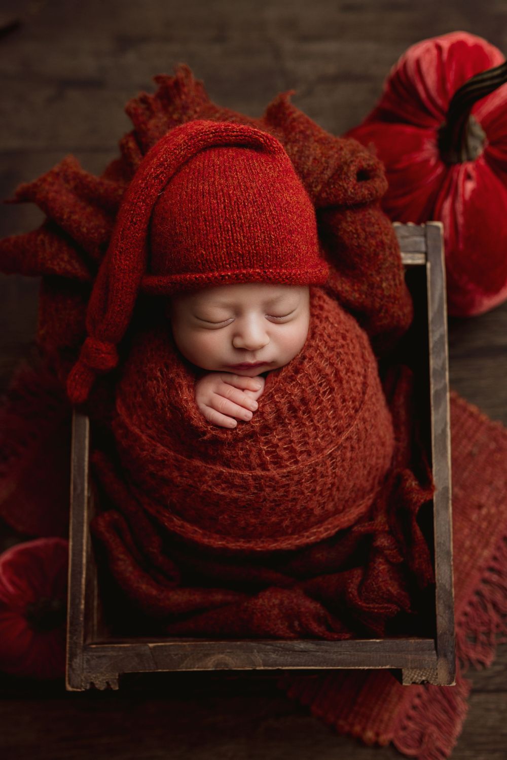 A sleeping baby wrapped in cozy deep red knit blankets and hat rests peacefully in a wooden box.