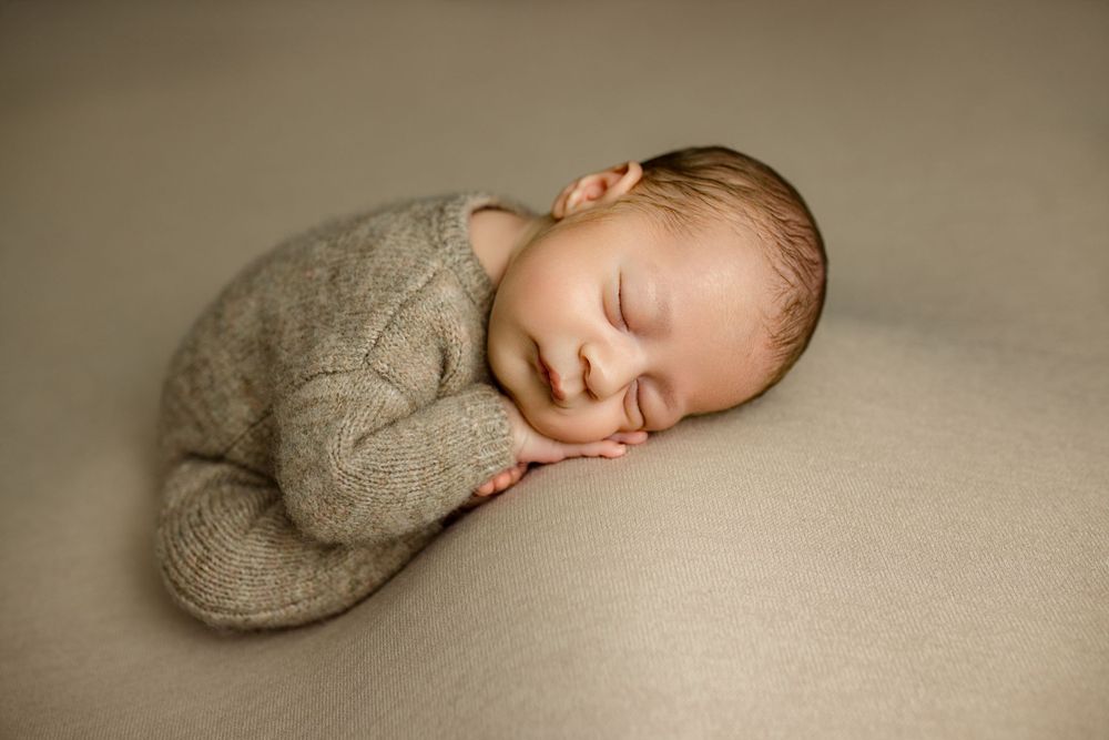 Sleeping newborn in soft gray outfit curled up on beige backdrop during professional photo session.