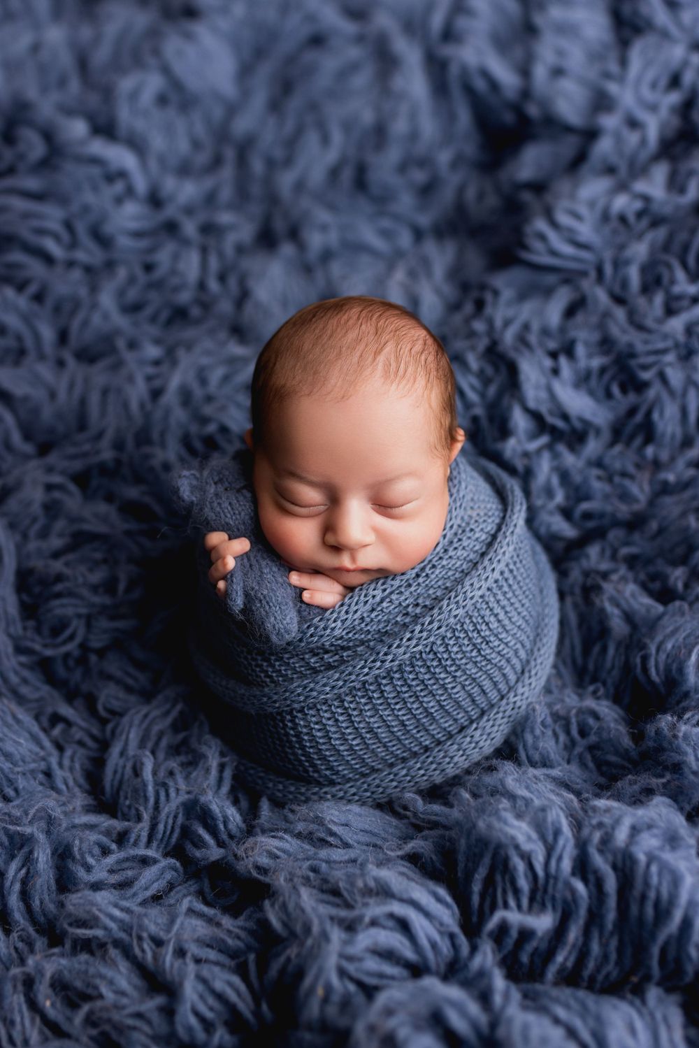 Newborn baby peacefully sleeping while wrapped in a cozy gray knit blanket on textured blue fabric.