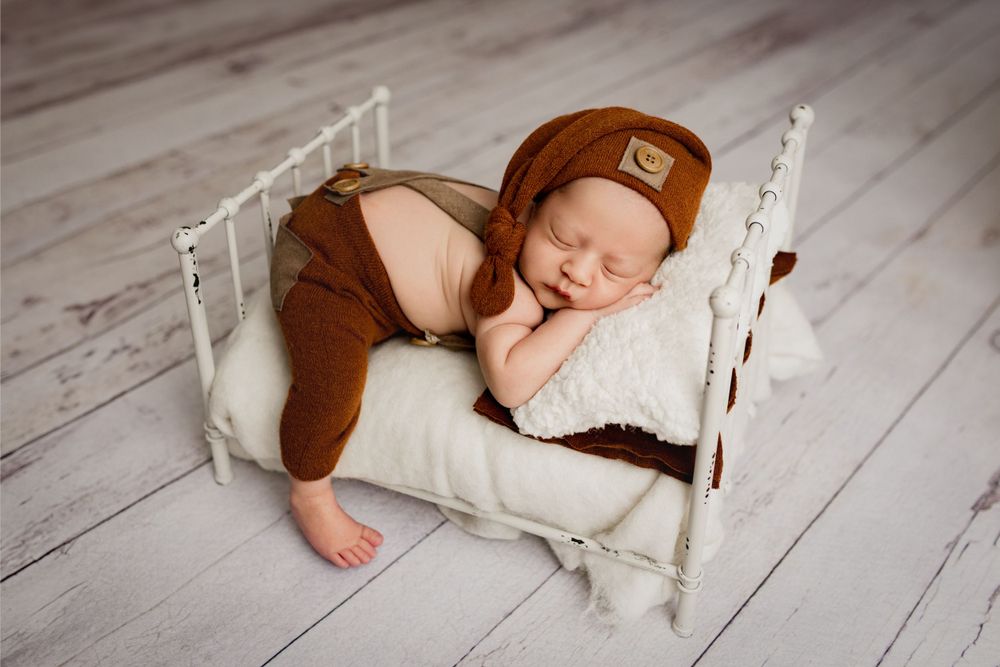 Sleeping baby wearing brown outfit rests on white pillow in rustic wooden bed prop on gray floor.