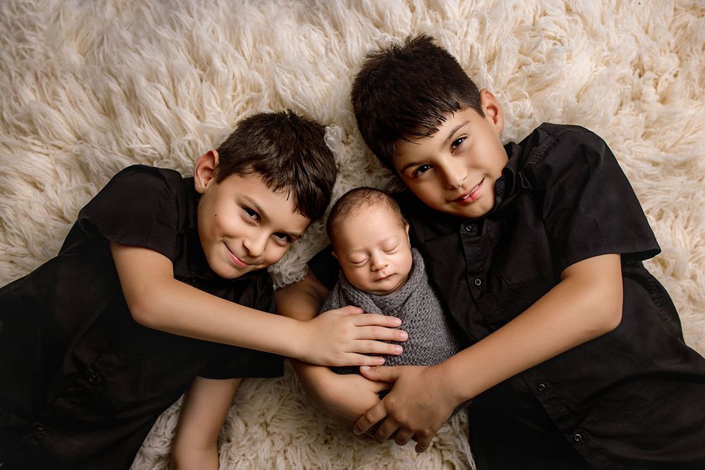 Three siblings wearing black clothing laying together on a cream colored fluffy rug.