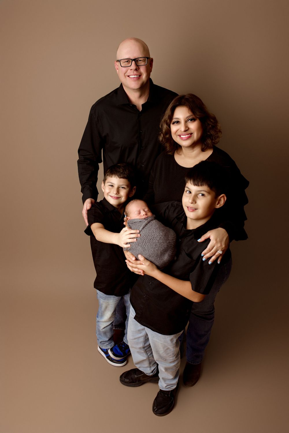 A family of five wearing coordinating black outfits poses together for a studio portrait against a brown backdrop.