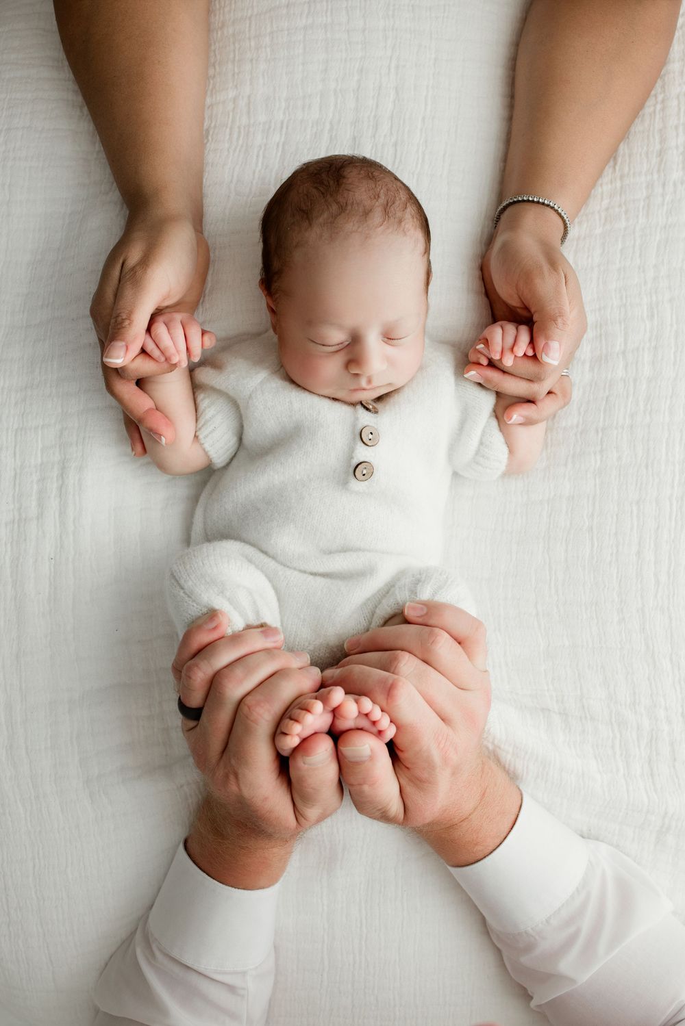 A sleeping baby in white clothing lies on light fabric while gentle hands hold their tiny fingers and toes.