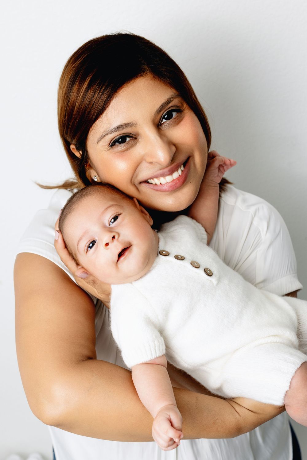 A tender moment captured between a parent wearing white and their young baby in matching white clothing against a light background.