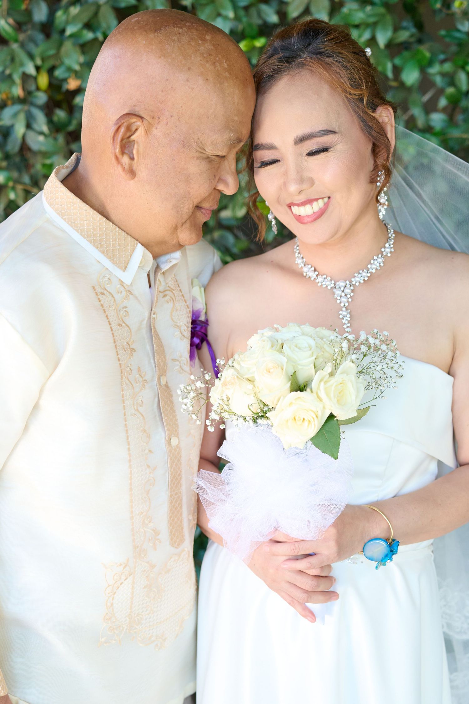 A bride in a white wedding dress and groom in traditional Filipino barong standing against a marbled blue backdrop.