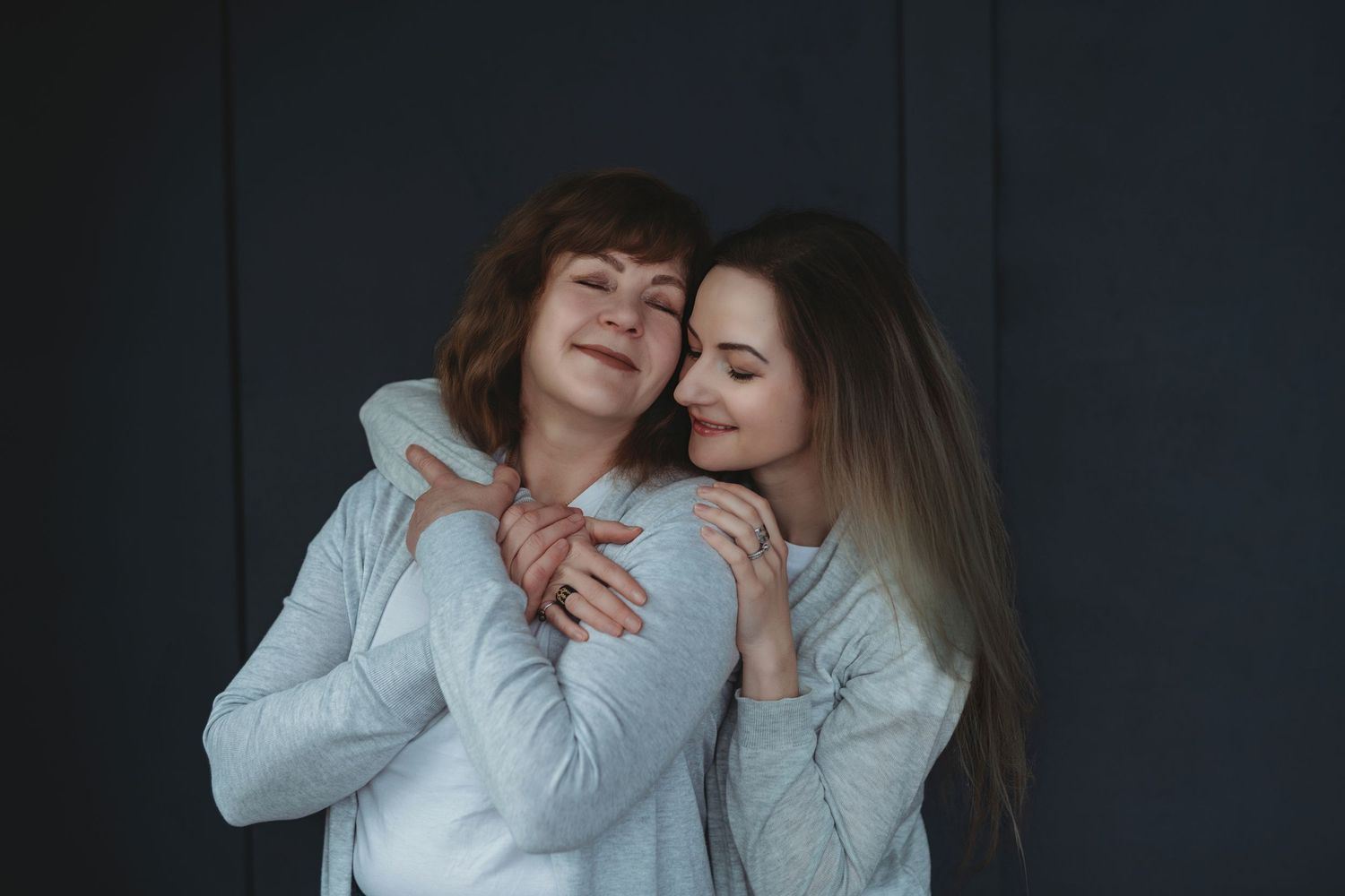 Two women in white sweaters laughing and embracing against a dark background.