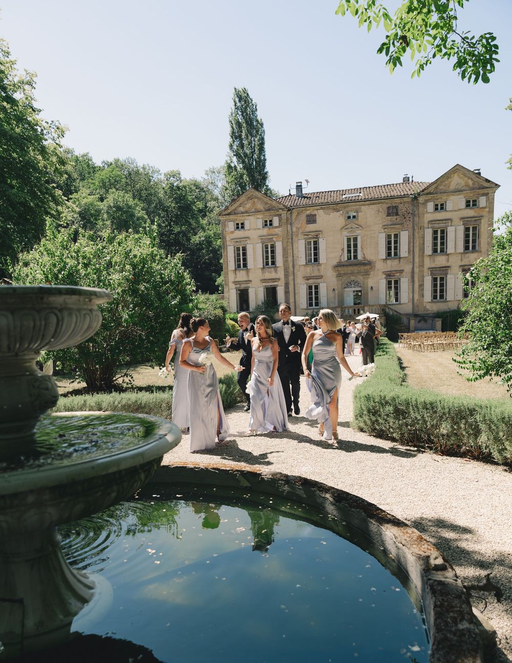 Wedding party walks past a stone fountain in lush gardens of a grand French chateau on a sunny day.