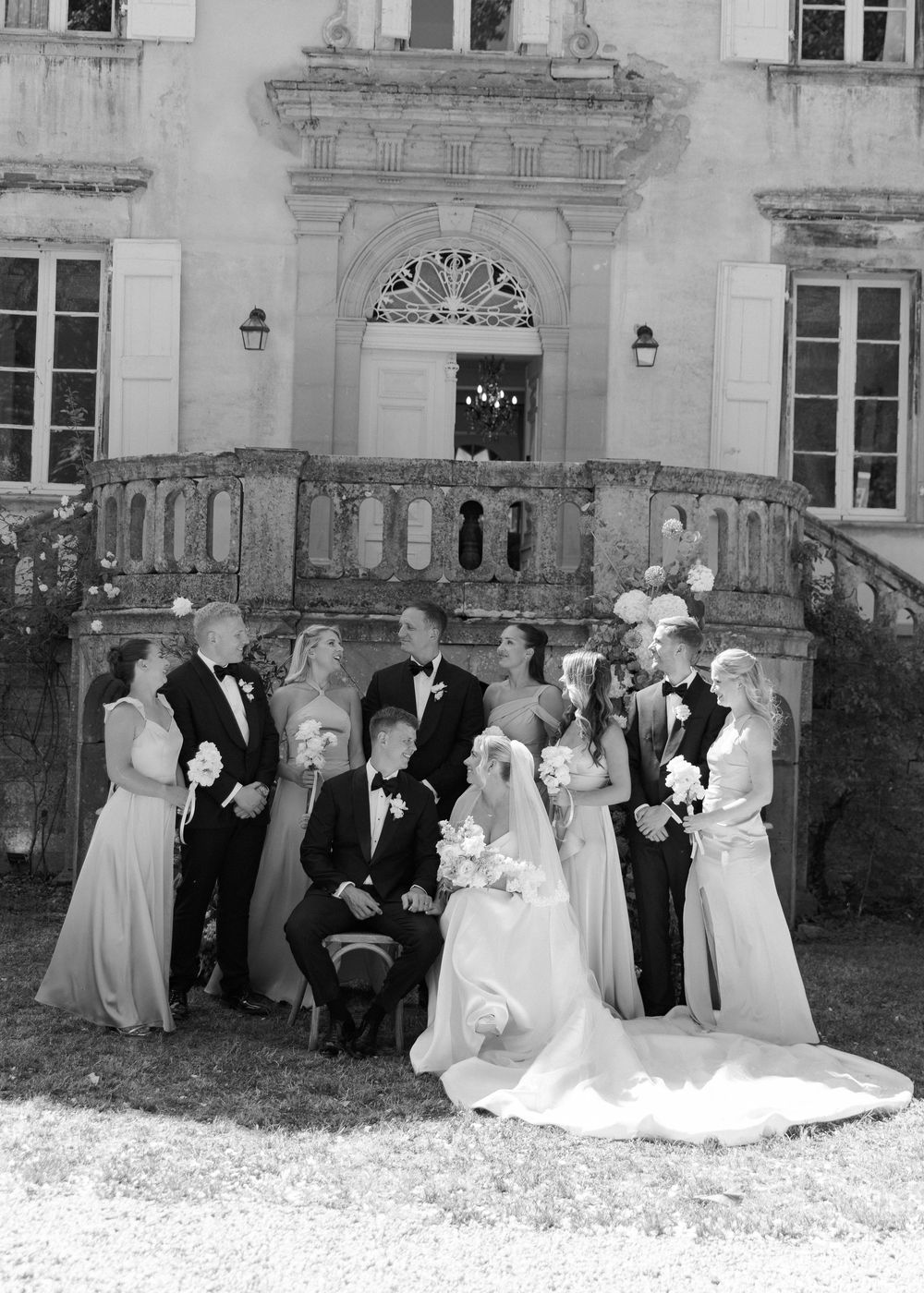 Black and white wedding party photo in front of elegant chateau with grand staircase.