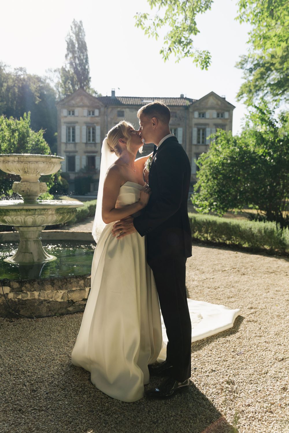 Bride and groom share a romantic embrace near a stone fountain in a lush garden with an elegant mansion backdrop.