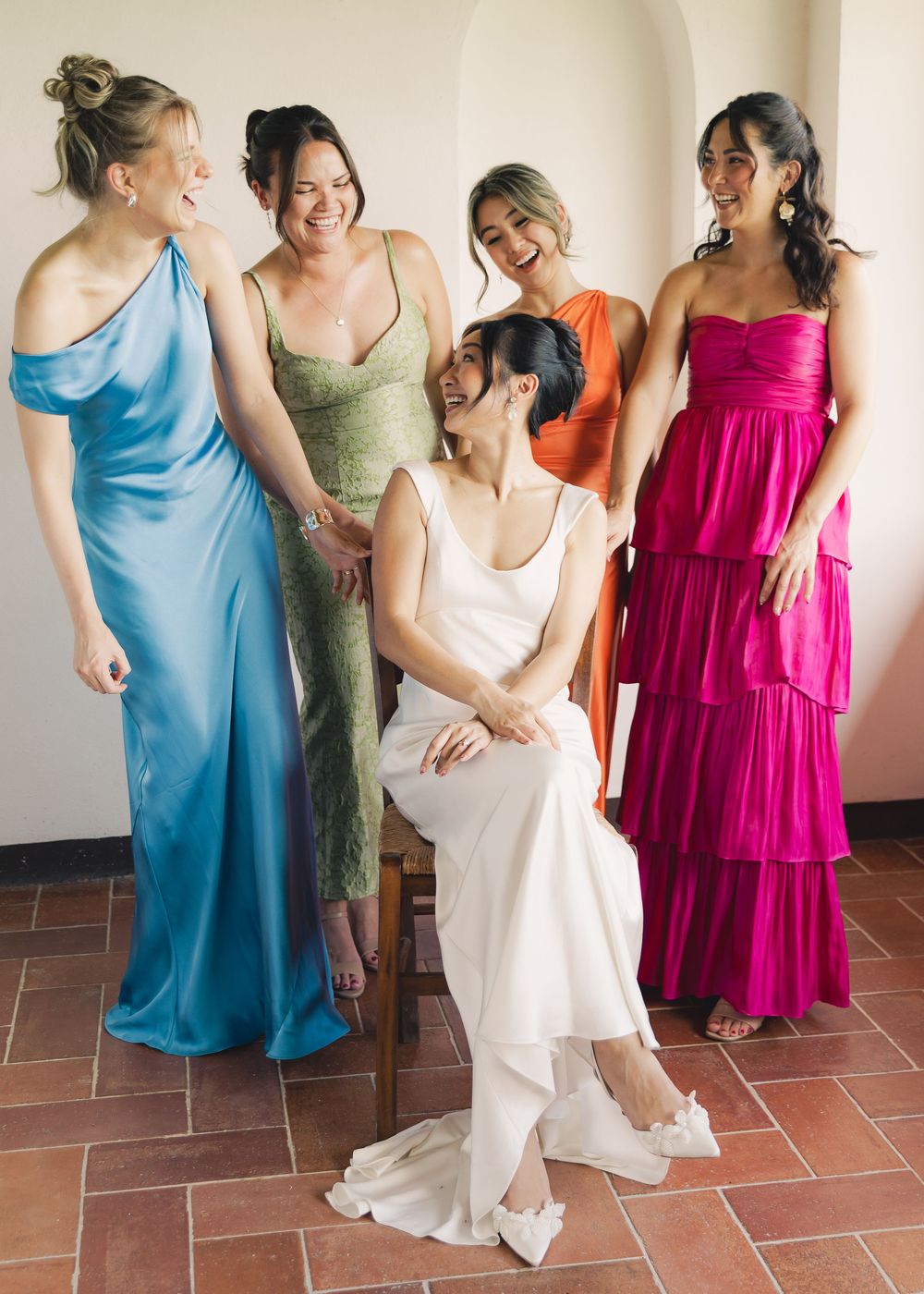 Bride in white gown seated surrounded by four bridesmaids in colorful dresses, smiling together indoors.