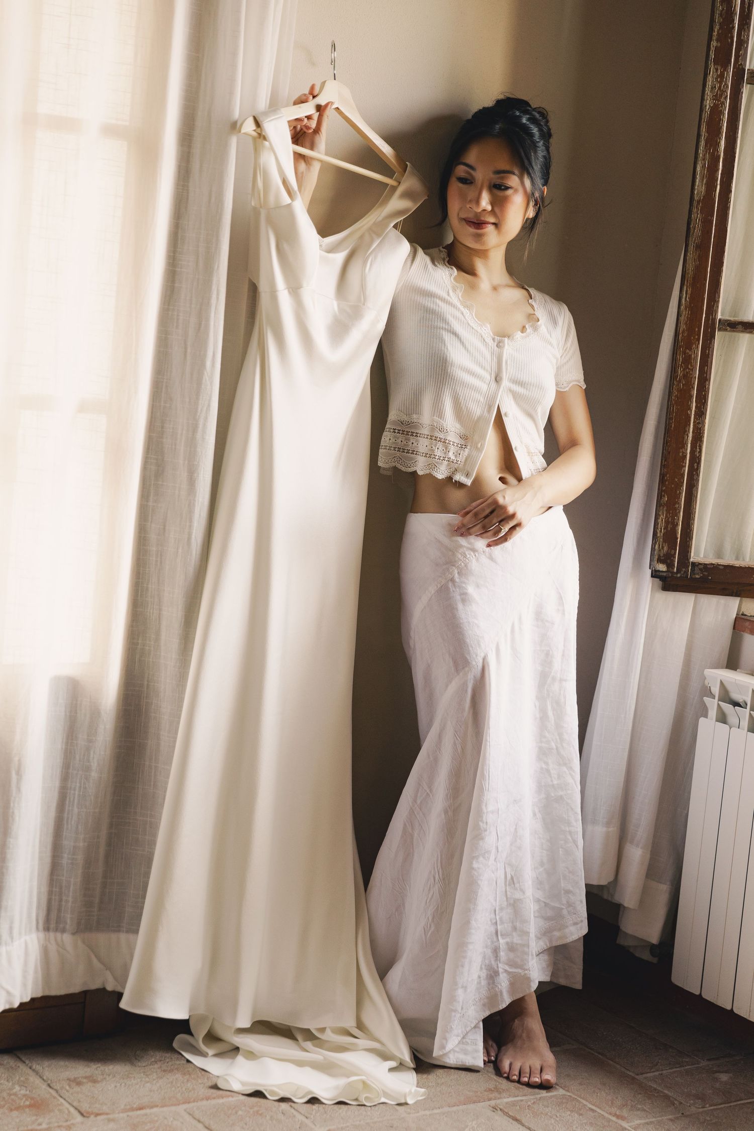 Bride holding wedding dress on hanger in white two-piece outfit, standing by window in elegant room.