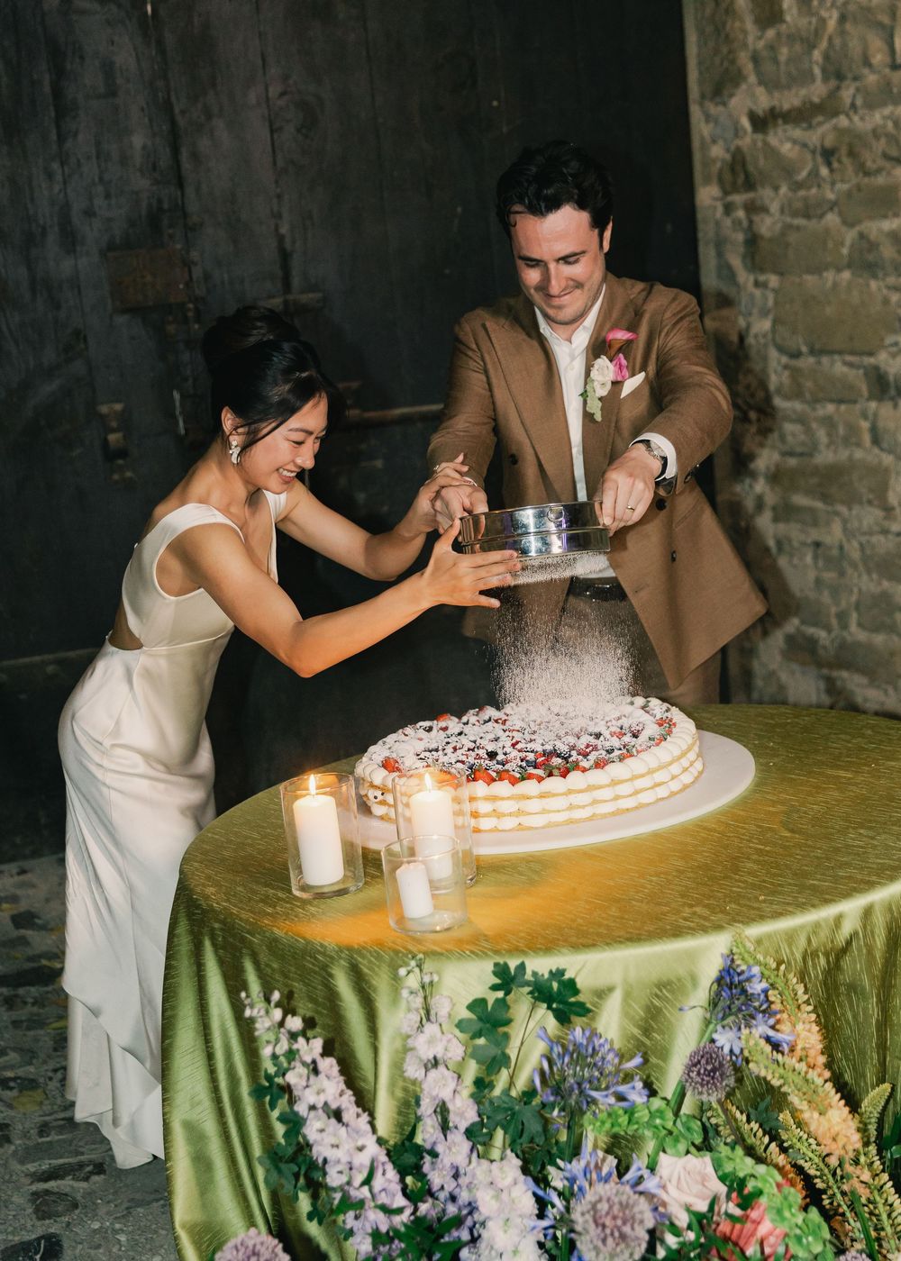 Bride and groom dusting powdered sugar on a white berry wedding cake at an elegant candlelit reception.