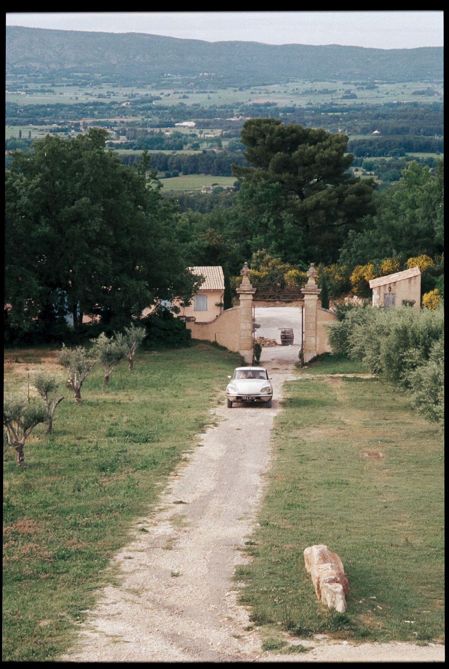White car drives down rural gravel path through ornate stone gate with scenic valley landscape beyond.