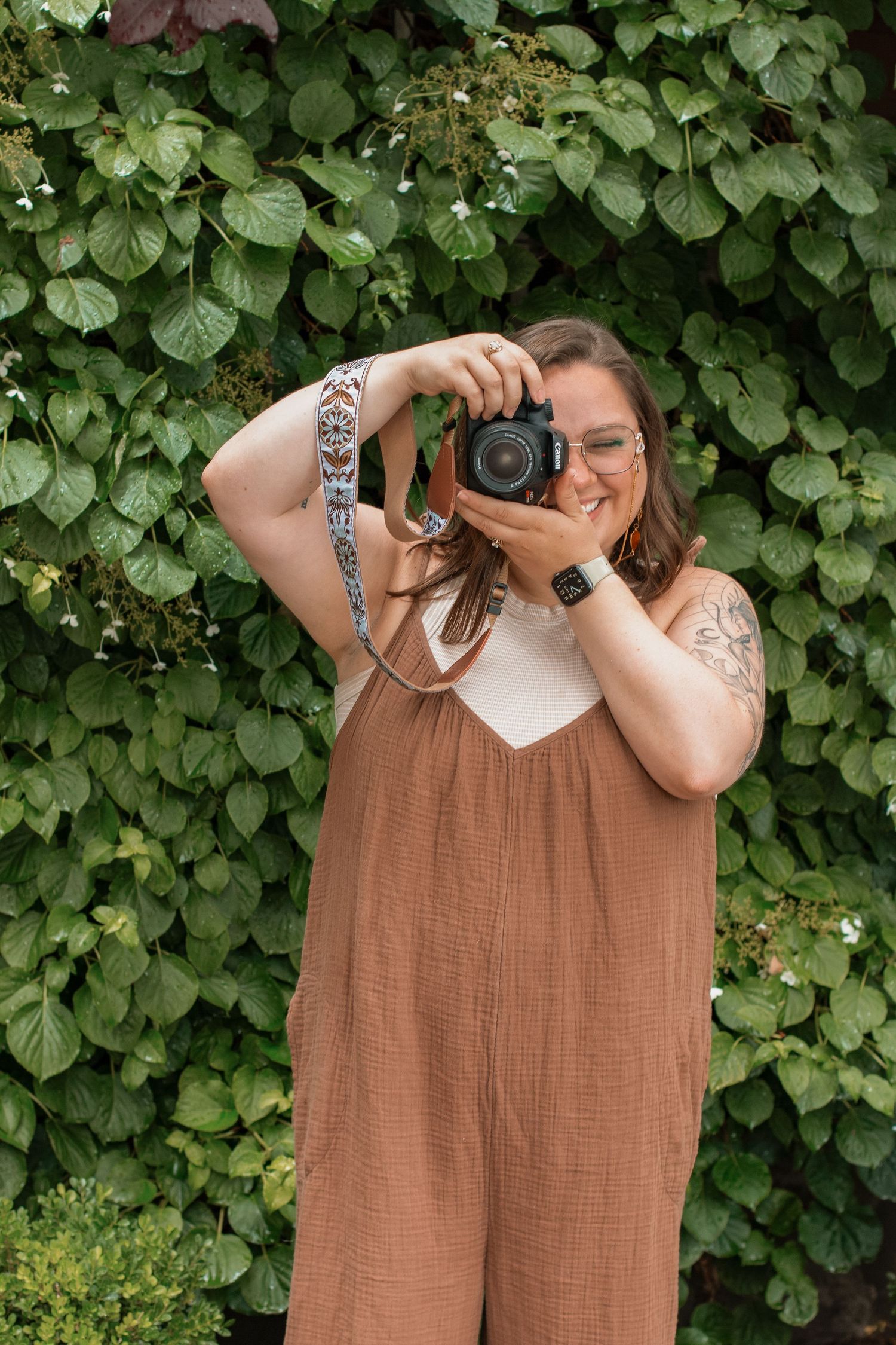 A photographer wearing a brown jumpsuit takes a picture with a DSLR camera against a leafy green backdrop.