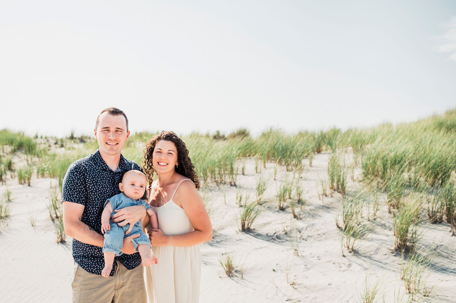 A family standing together and smiling on a sandy beach with beach grass in the background.