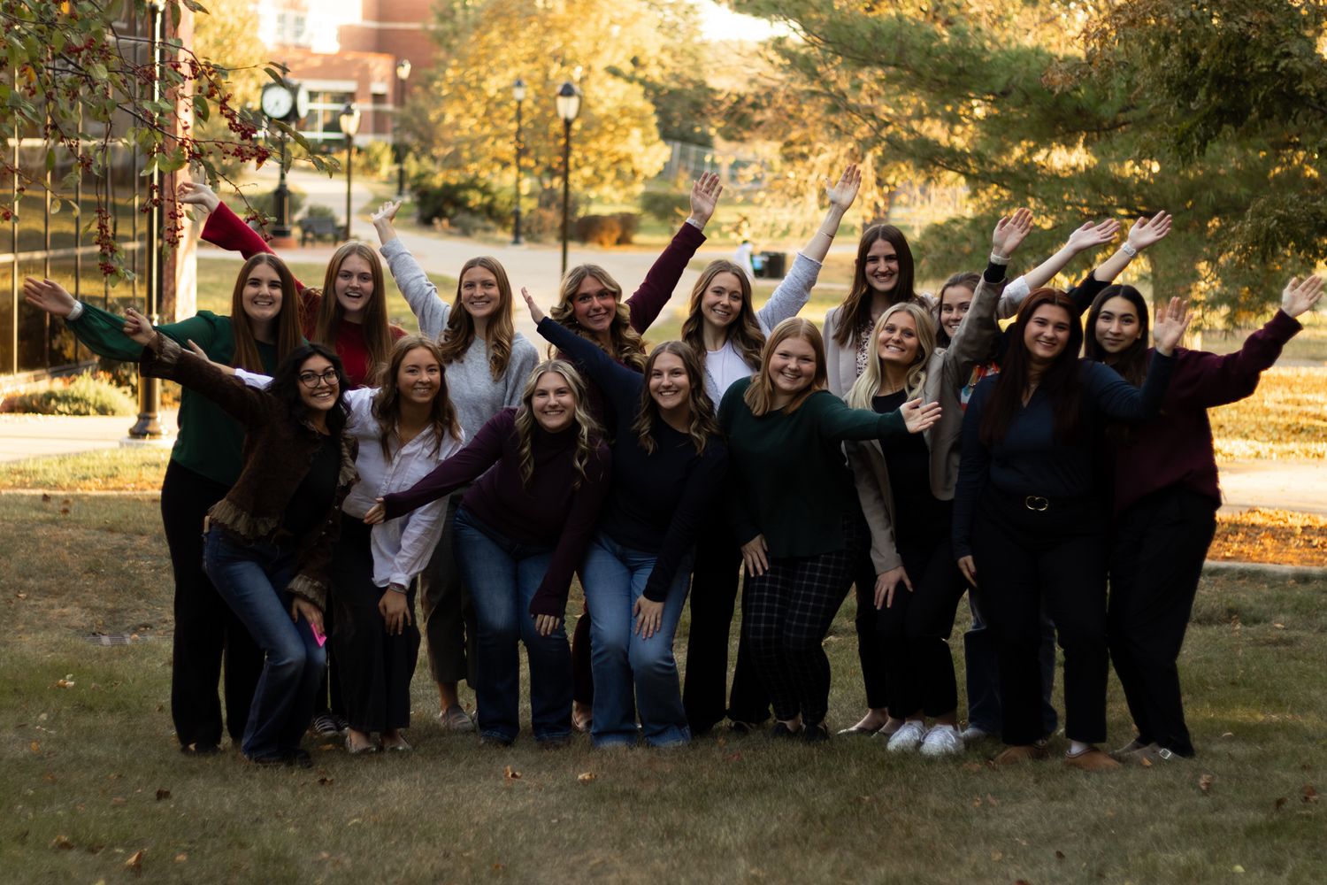 A group of college students in casual attire pose together outdoors on an autumn day with golden trees in the background.