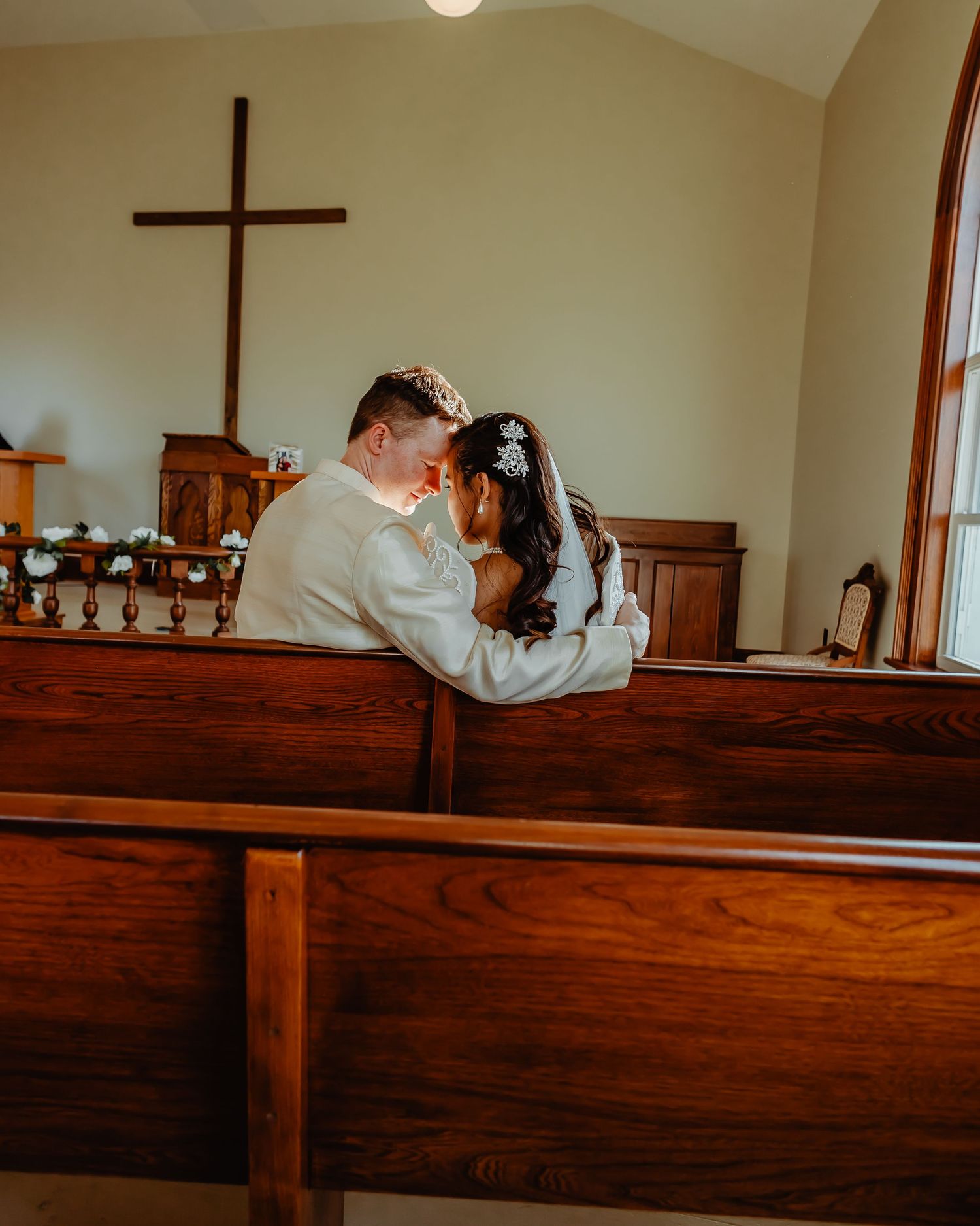 Couple shares an intimate moment while sitting together on wooden church pew near cross and stained glass window.