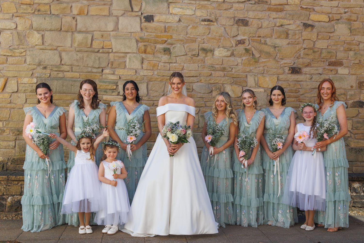 Wedding party in sage green and white dresses posing in front of a rustic stone wall.