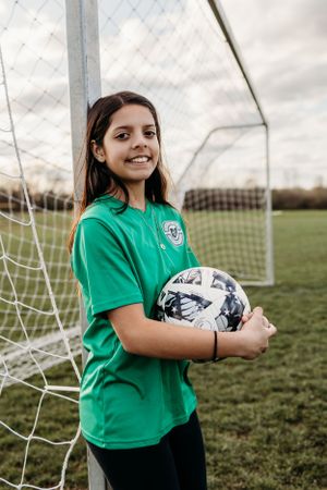 Young soccer player in green jersey holds black and white ball while standing by goal post on grass field.