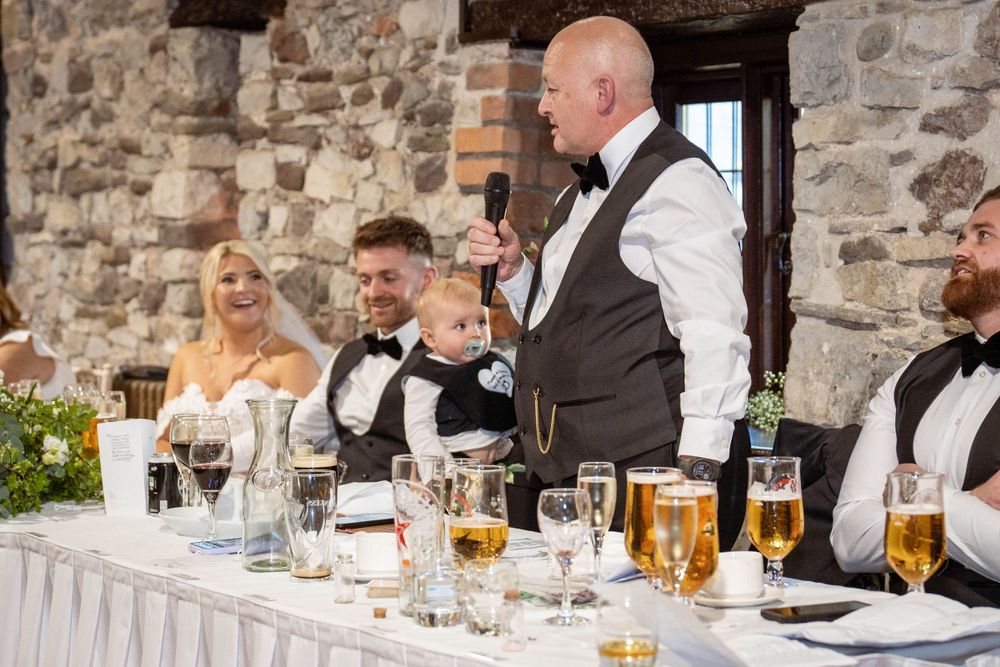 Wedding party members seated at head table during reception celebrations in historic stone building.
