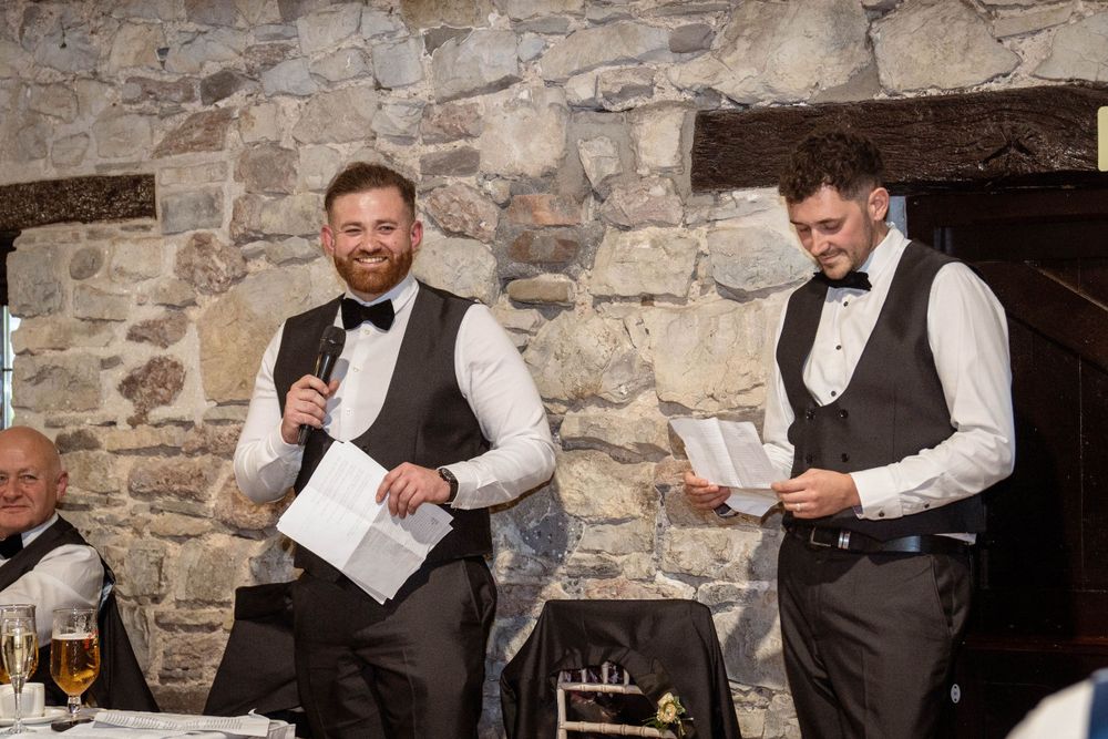 Groomsmen in formal attire share drinks and smiles at a rustic stone wedding venue.