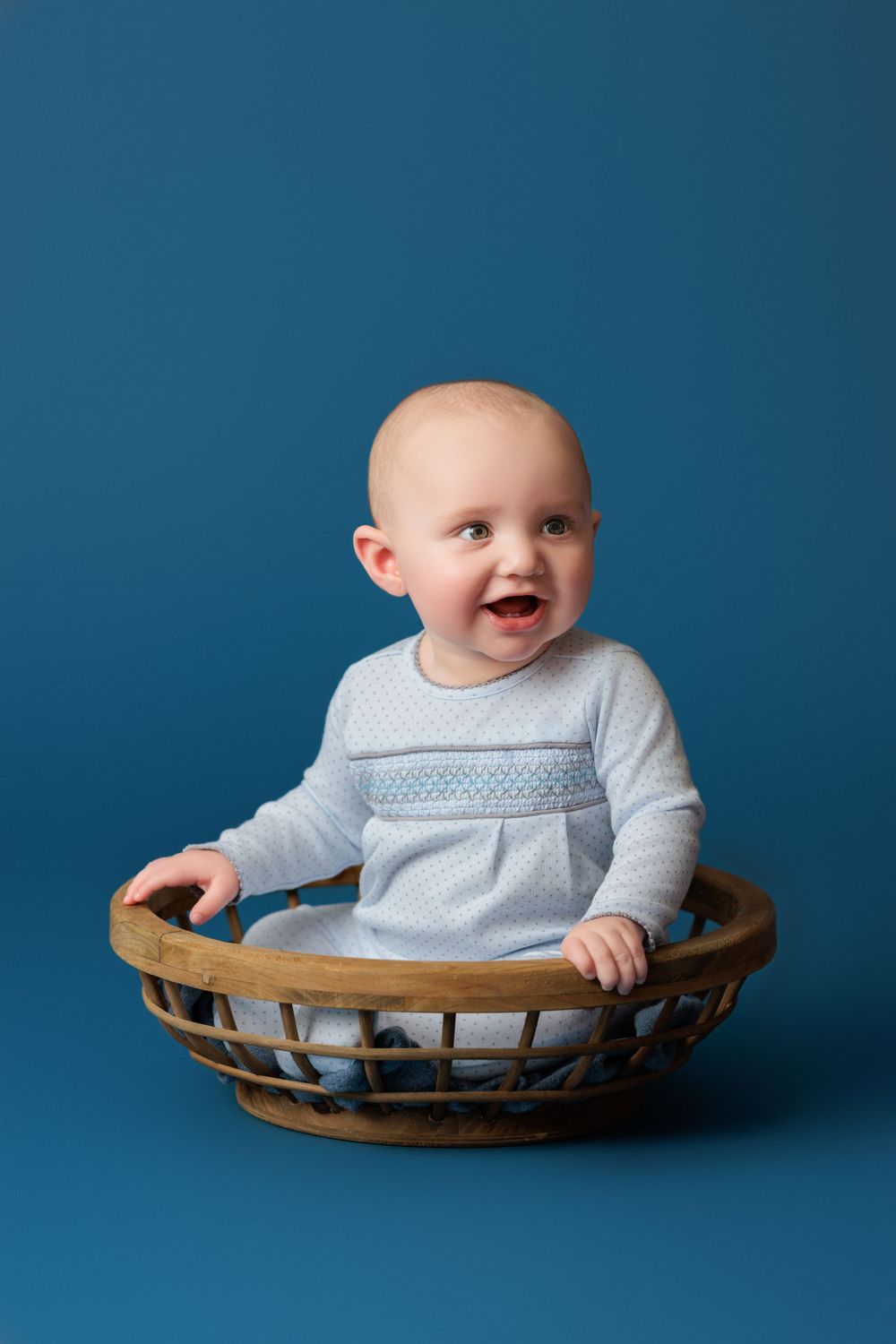 Baby in light blue outfit sitting inside vintage wicker basket against blue background.