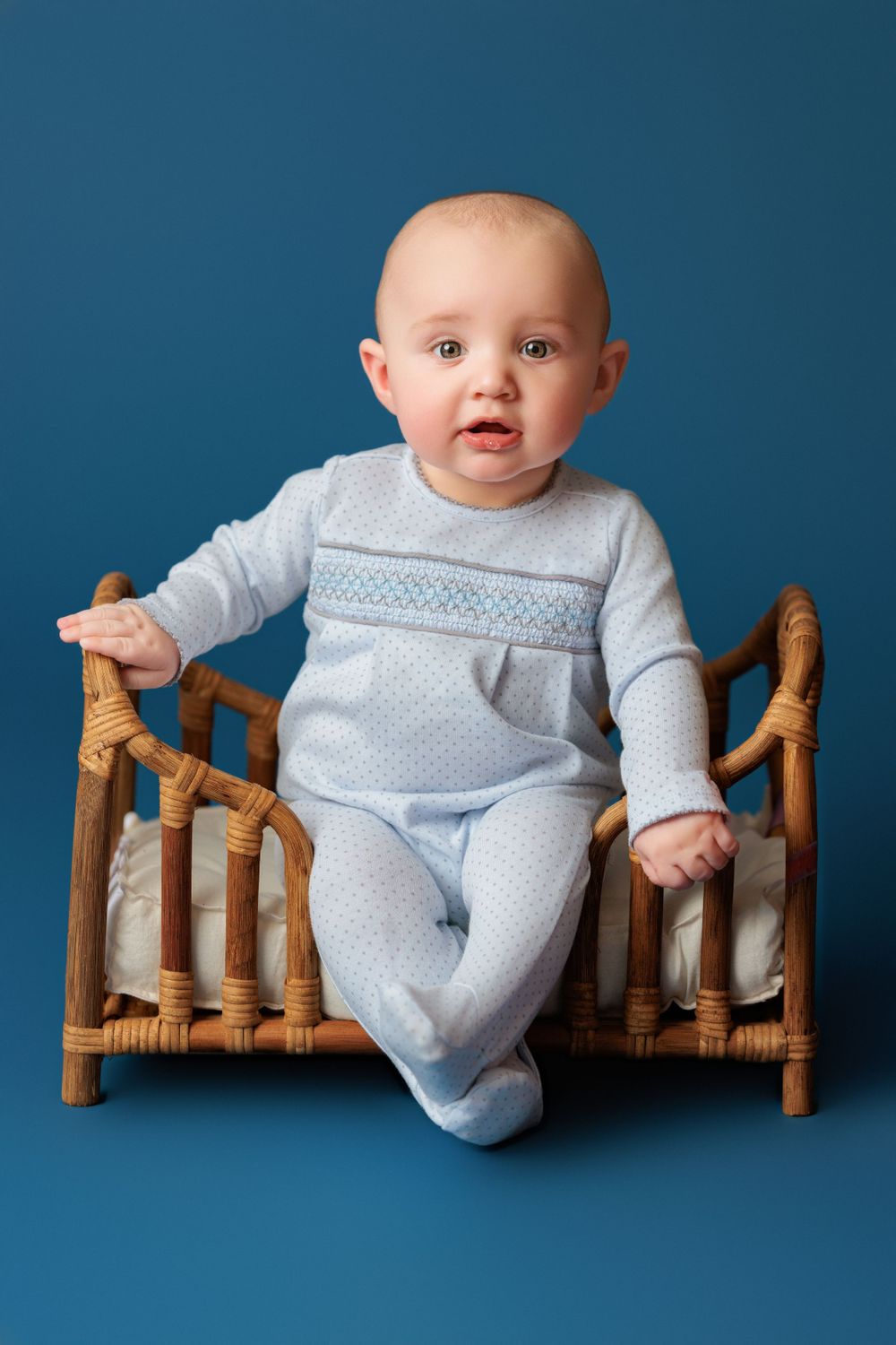 Baby in light blue polka dot outfit sits on wooden rattan bench against blue backdrop.
