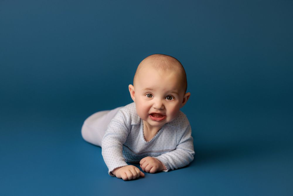 Baby in grey outfit lying on tummy against blue studio background.