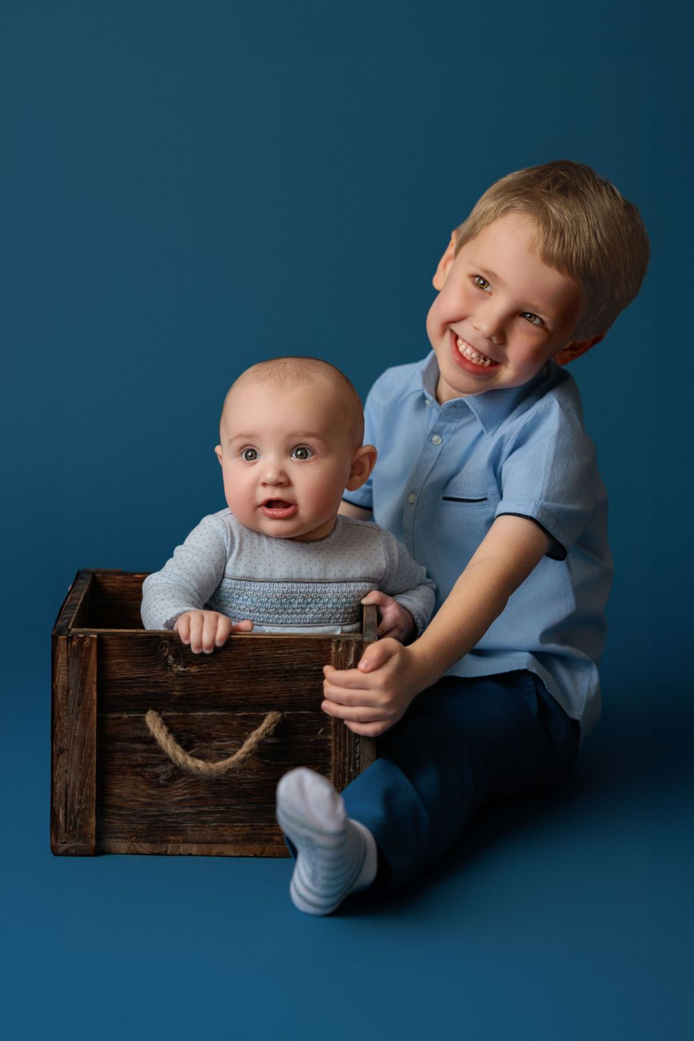 Two siblings pose together with a wooden crate against a blue backdrop during a studio photoshoot.