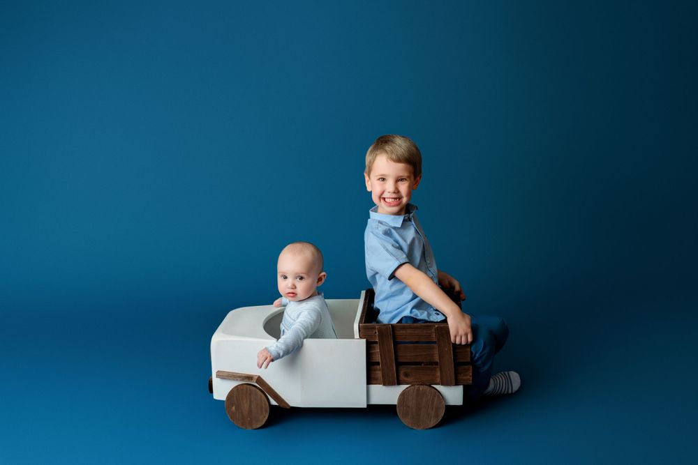 Two children pose on wooden toy trucks against a deep blue studio backdrop.