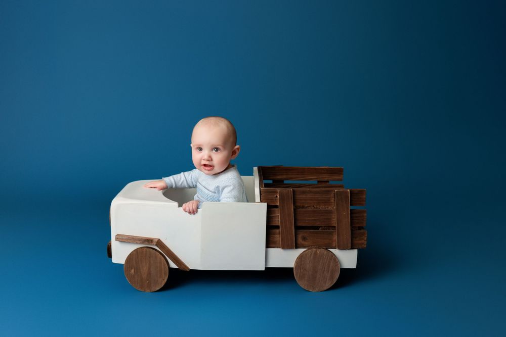 Baby sitting in wooden toy truck against blue background during professional studio photo session.