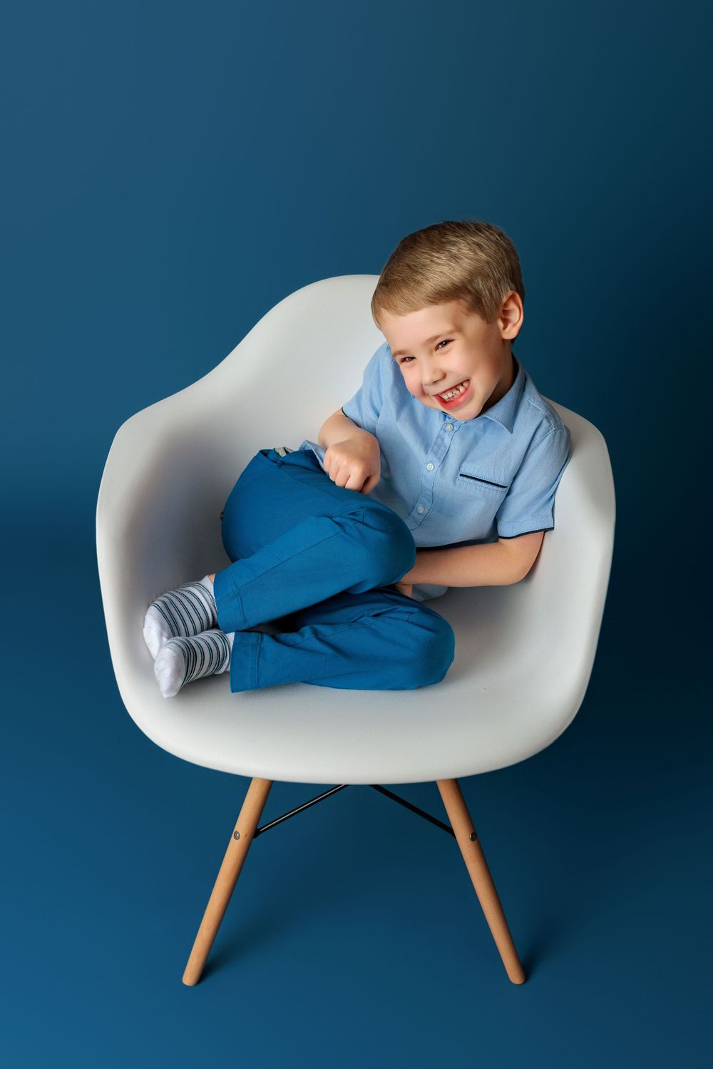 Child in light blue shirt and navy pants sits curled up in a modern white chair against blue background.