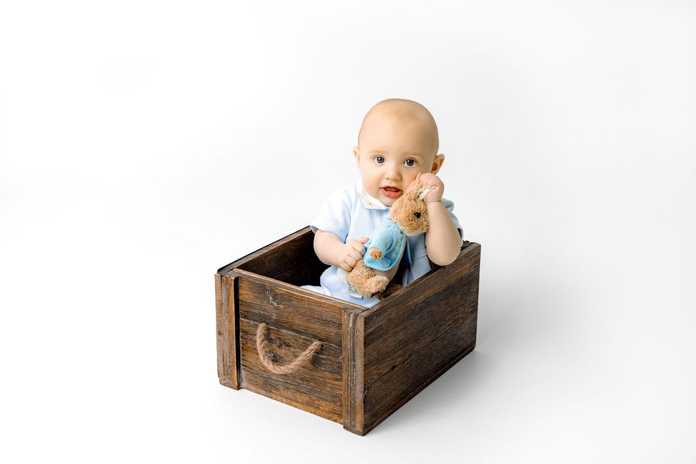 Baby in light blue outfit sits in a rustic wooden box with etched bird detail during photo session.