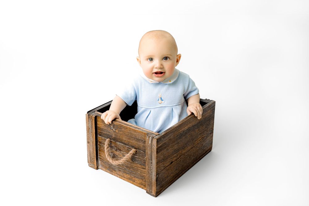 A small child wearing light blue sits in a rustic wooden crate against a white background.
