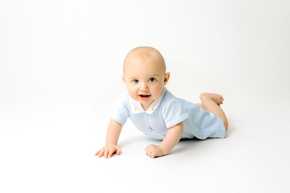Baby in light blue onesie crawls on white background during professional portrait session.