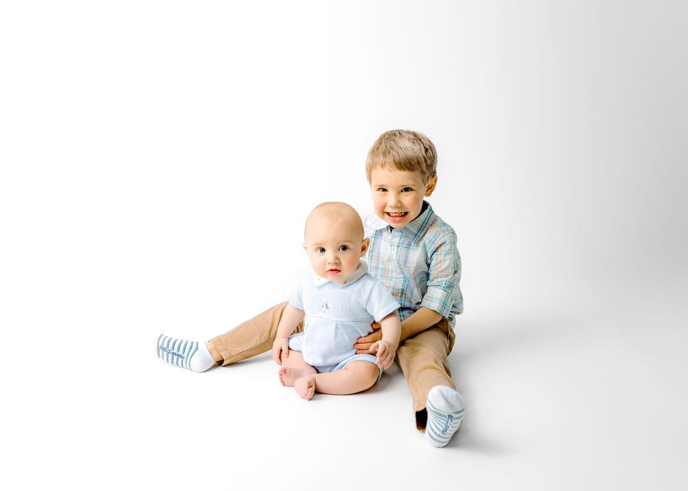 Two young children sit together on white background wearing casual outfits and striped socks.