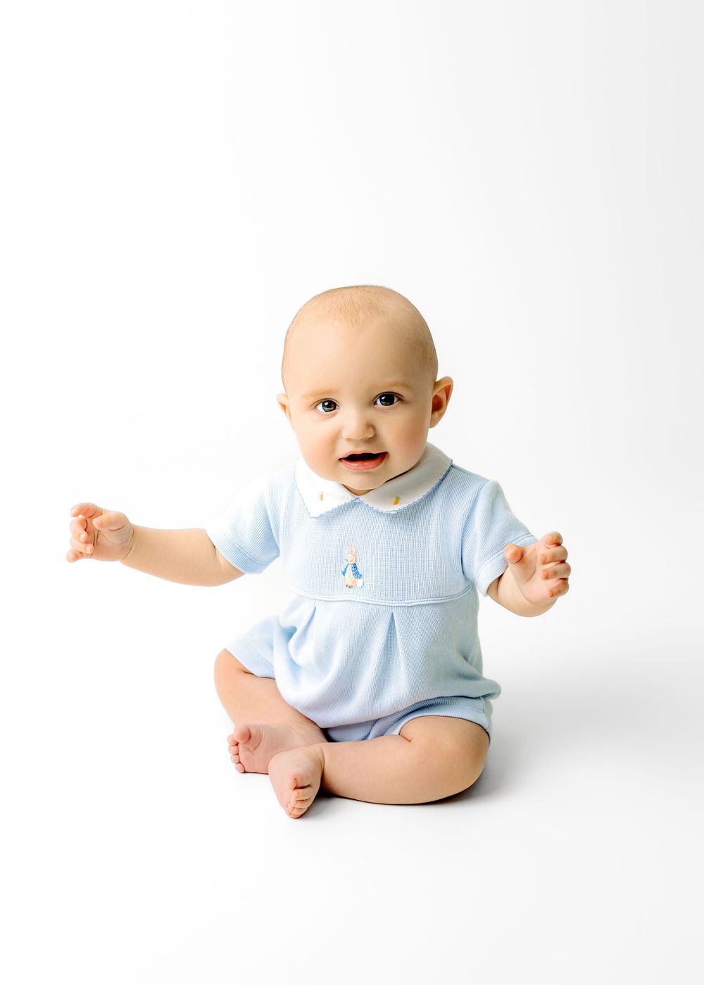 Baby in light blue romper sitting on white background with arms outstretched.