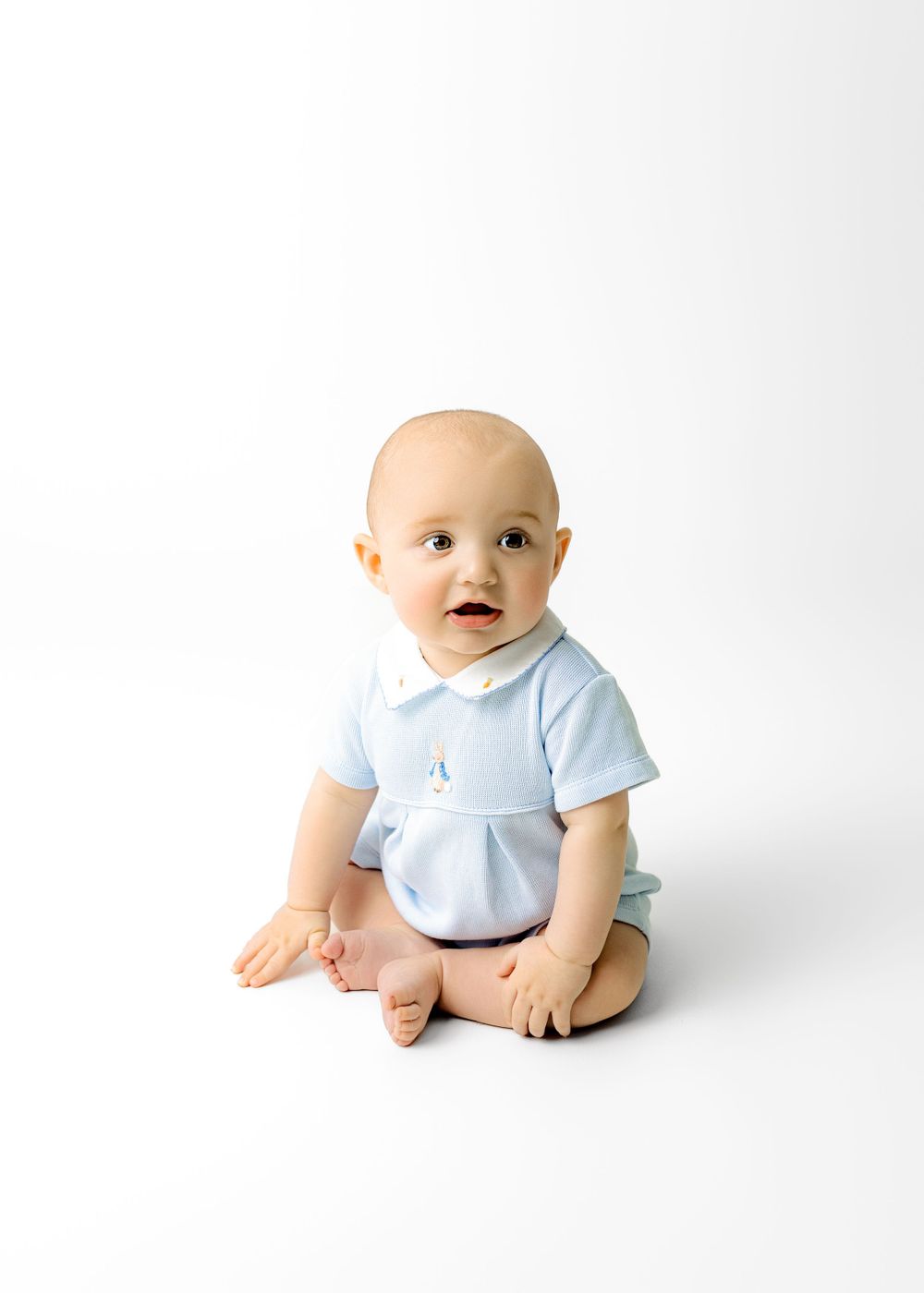 Baby wearing light blue outfit sitting on white backdrop during studio photoshoot.