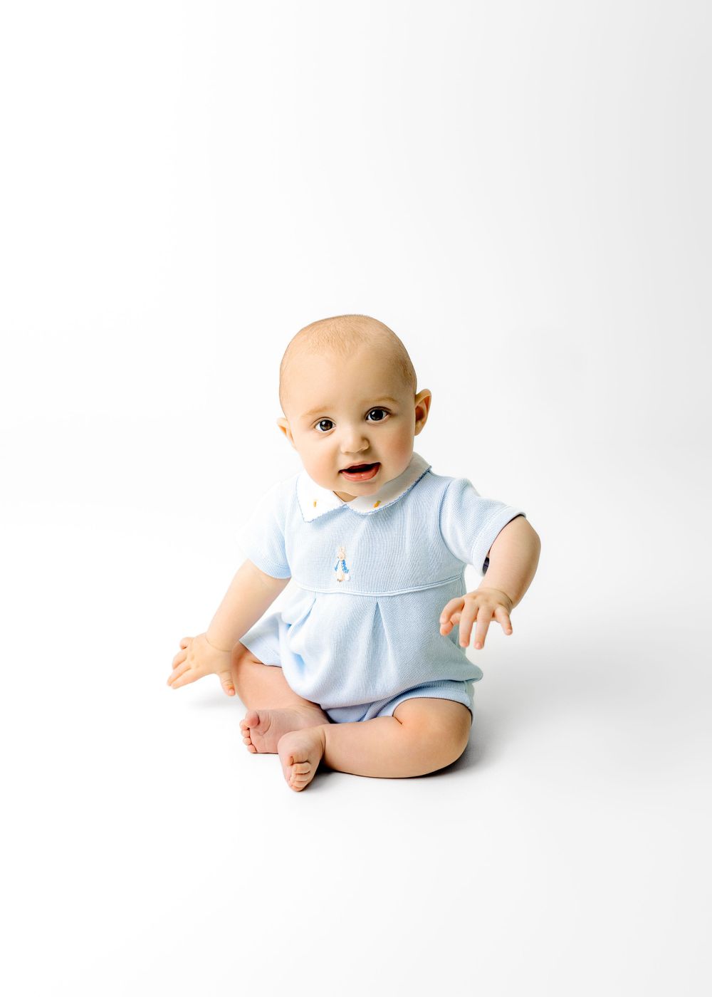 Baby wearing light blue romper sits on white background with happy expression and bare feet.