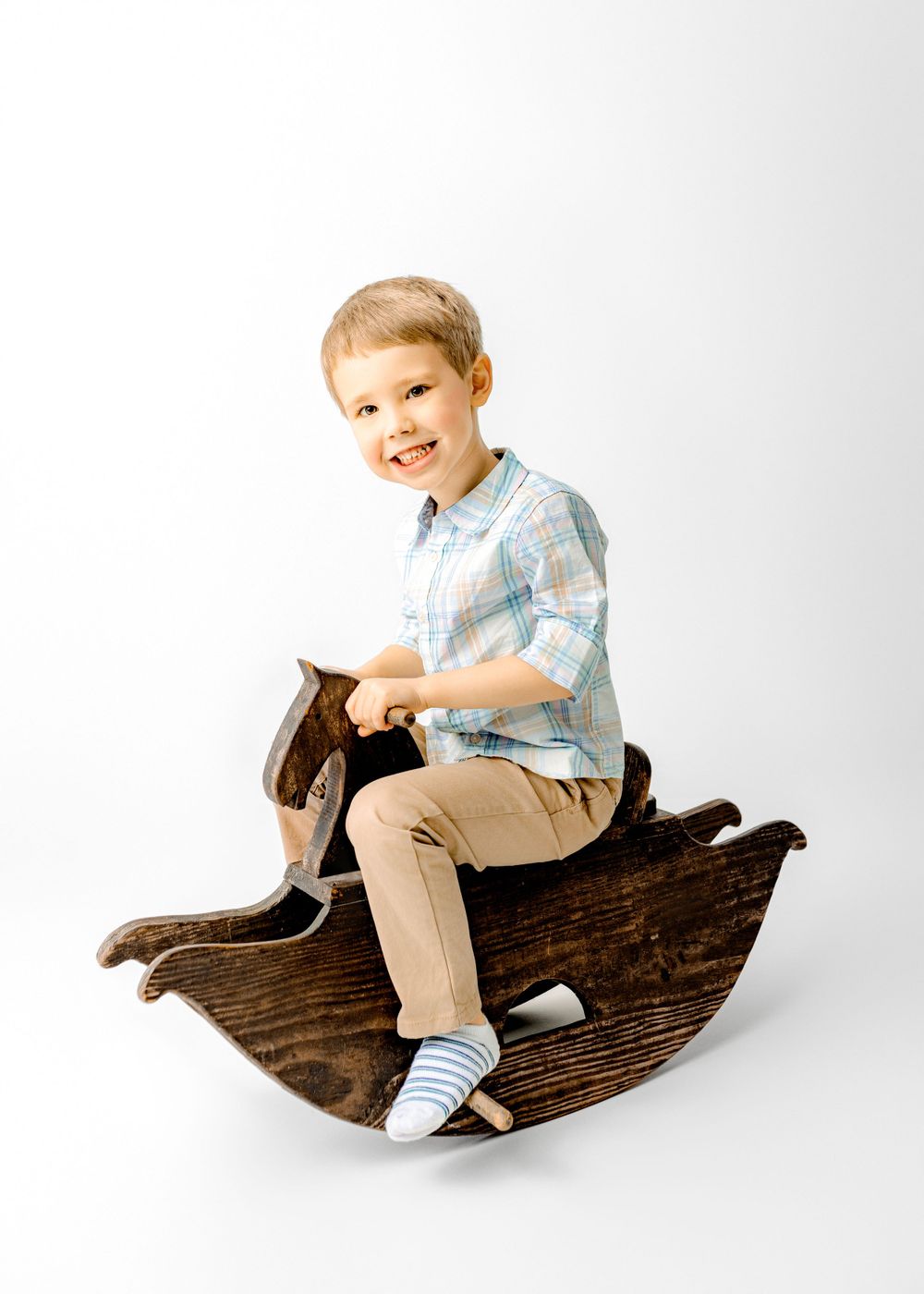 Smiling child sits on wooden rocking horse toy against white background during photo session.