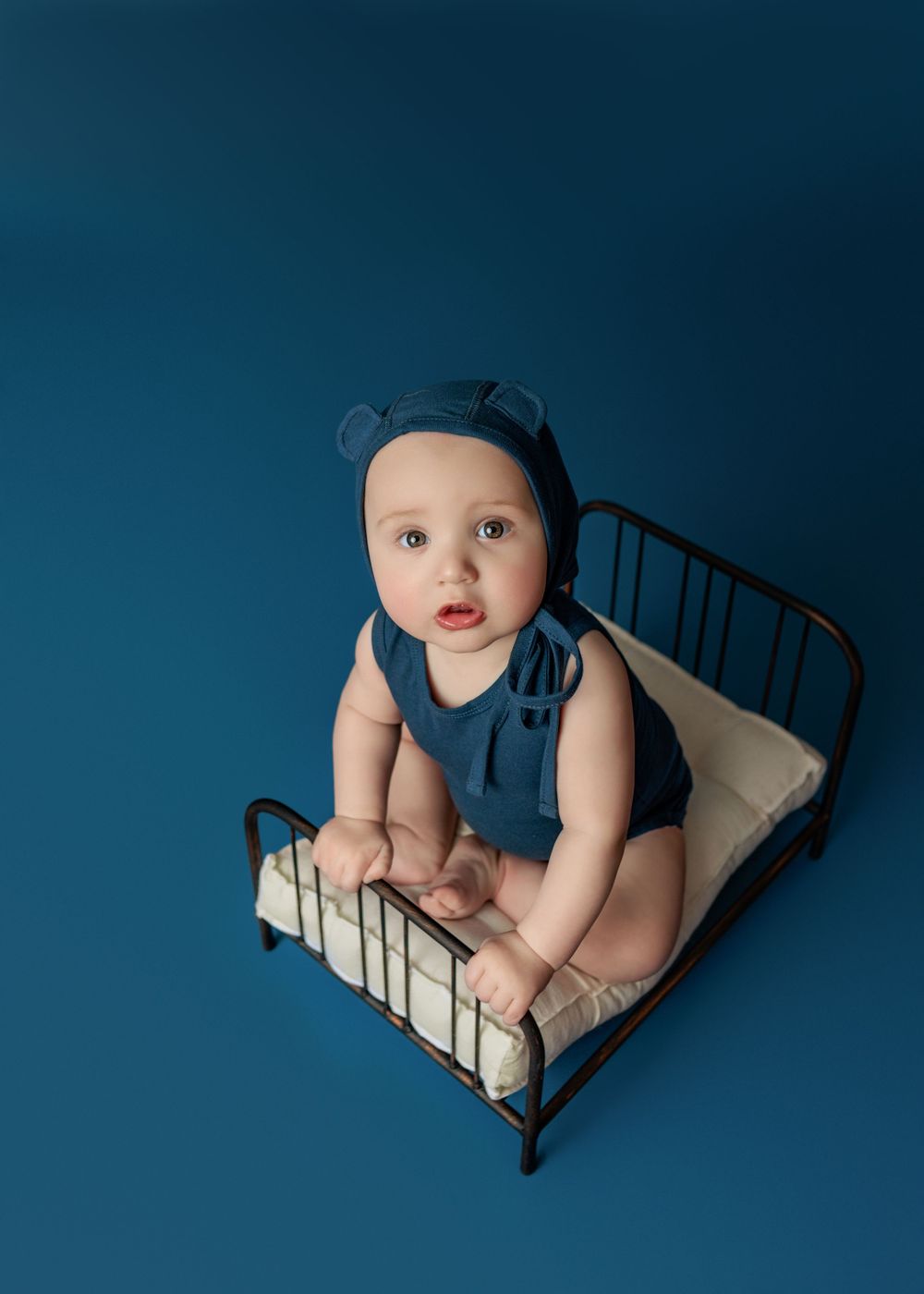 Baby wearing navy outfit sits on small iron bed with white bedding against blue backdrop.