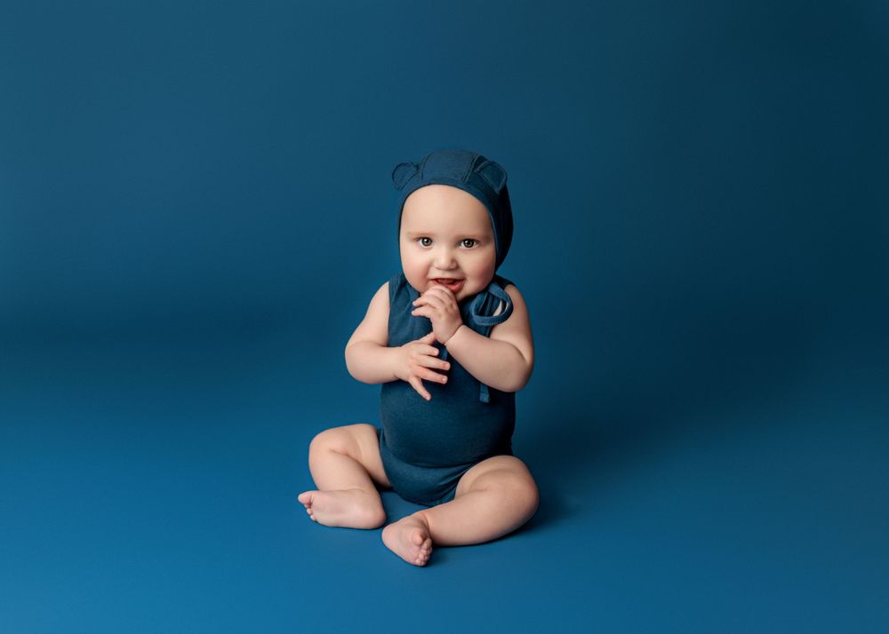 Baby in navy blue outfit sits on blue background with hands clasped.