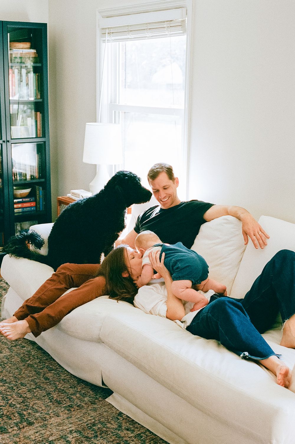 Family relaxing together with dog on white couch in sunlit room.