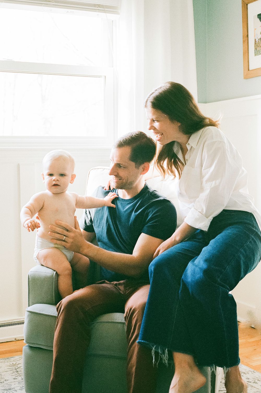 A family sits together in a cozy living room by a bright window, enjoying a tender moment with their baby.