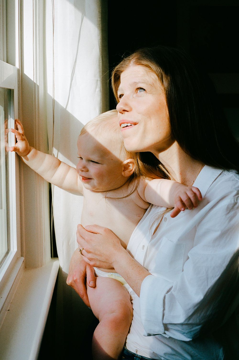 Parent and baby looking out sunny window together.